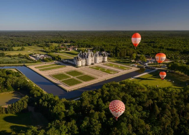 Vue aérienne du domaine national de Chambord avec en premier plan 3 montgolfières, puis les jardins à la française et au milieu de la photo le château.