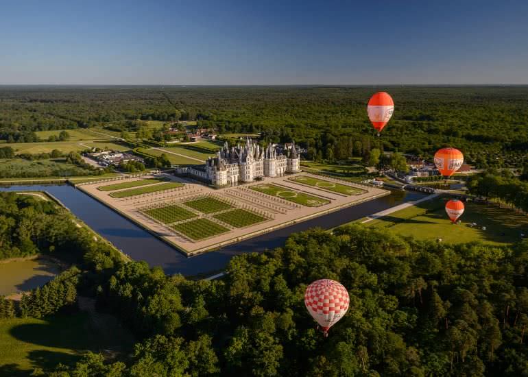 Vue aérienne du domaine national de Chambord avec en premier plan 3 montgolfières, puis les jardins à la française et au milieu de la photo le château.
