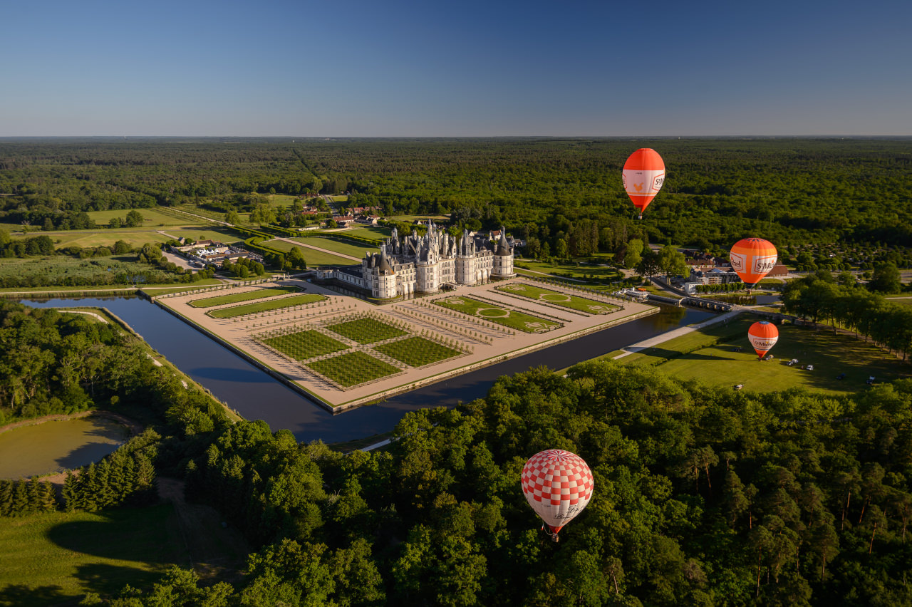 Vue aérienne du domaine national de Chambord avec en premier plan 3 montgolfières, puis les jardins à la française et au milieu de la photo le château.